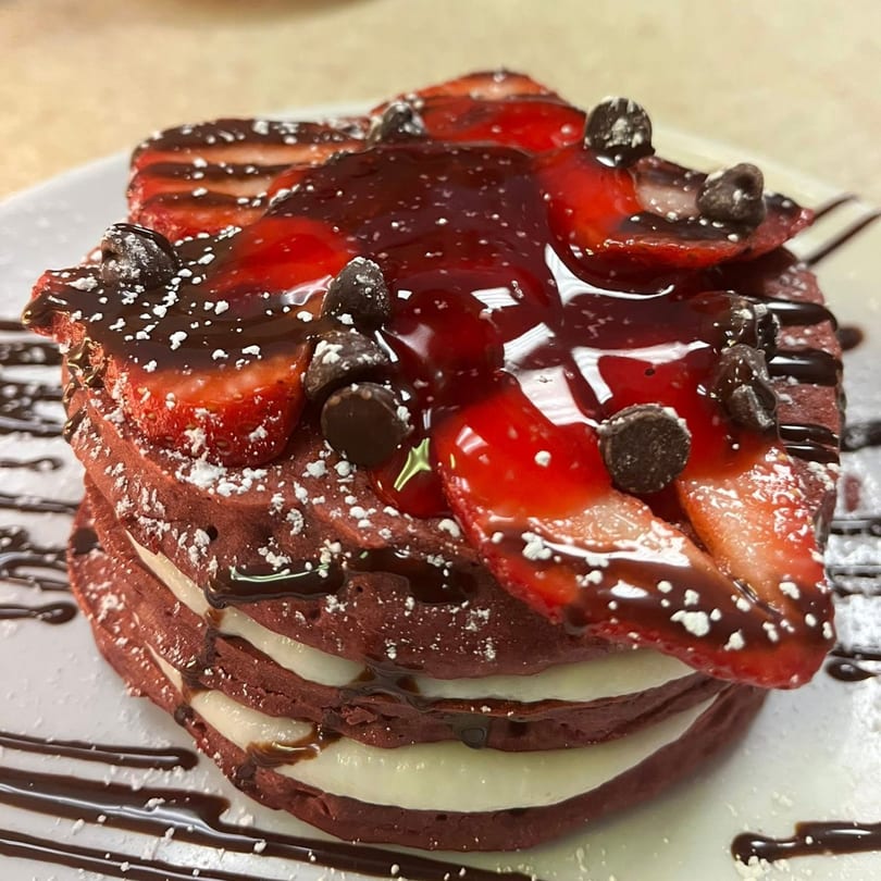 Red velvet pancakes with cream cheese filling, topped with strawberries, chocolate chips, and syrup.