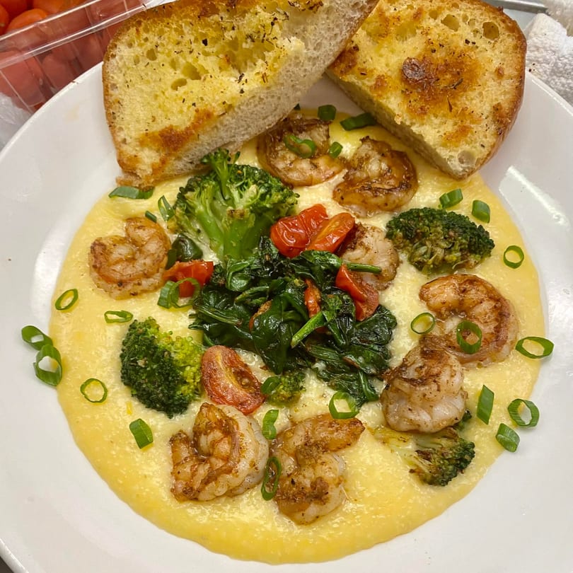Shrimp and grits with broccoli, spinach, tomatoes, and garlic bread on a white plate.