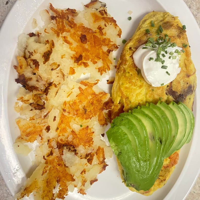 A fluffy omelet topped with avocado slices, sour cream, chives, and a side of crispy hash browns.