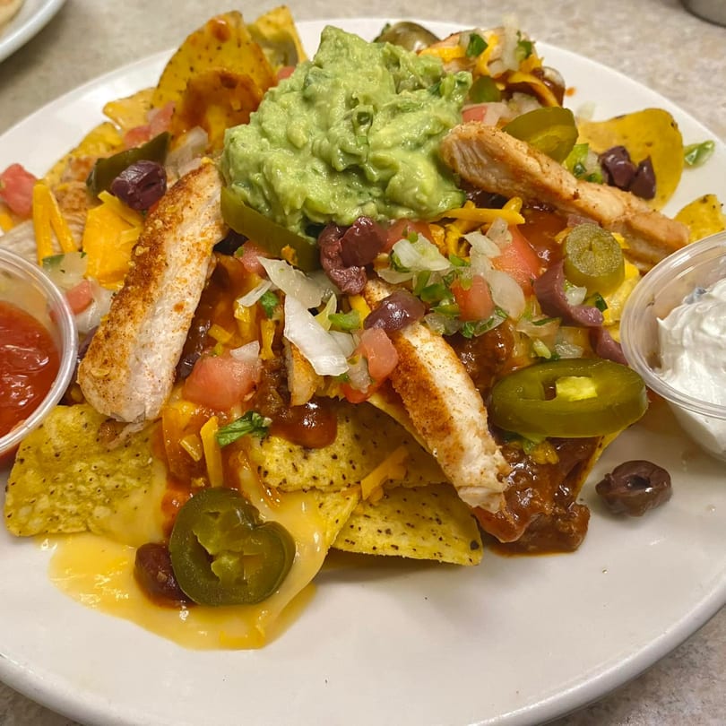 Chicken nachos with guacamole, salsa, sour cream, jalapeños, olives, and cheese on a white plate.